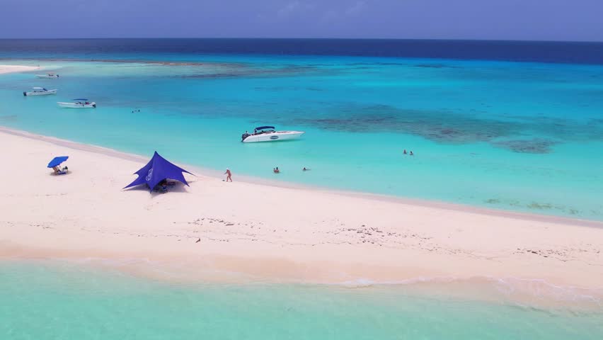 Zoom-out aerial of Cayo de Agua, Los Roques, Venezuela. Boats, people, and tent on white sand surrounded by turquoise Caribbean sea.