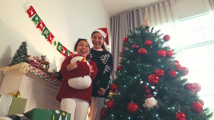 Two women—friends or mom and daughter—laugh in Santa hats by a Christmas tree with ornaments, lights, sparkle, ribbons, and presents. A joyful noel moment filled with warmth and togetherness.