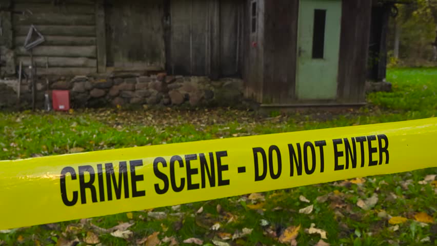 Yellow colored police crime scene forensic investigation tape ribbon in front of an old wooden traditional farm house or barn shed during a autumn day with fallen yellow leaves on the ground, bokeh.