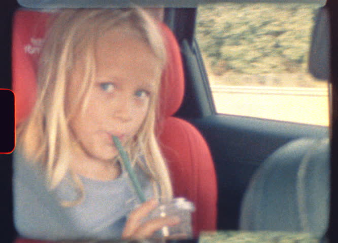 Blonde child sitting in the back seat of a moving car sipping cacao through a straw captured on vintage super 8 film