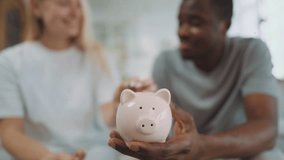 Closeup of funny ceramic piggy bank in hand of black man, woman putting coins. People save money for buying house and dreams realizing, family budget and finance, happy multiracial married couple - Powered by Shutterstock - Get 15% off with code: PIKWIZARD15