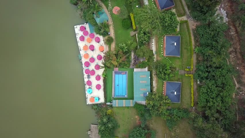 Top-down aerial descend over lakeside hotel at Lake Bunyonyi, Uganda. Pool, dock with umbrellas, cabins, and gardens blend into lush terrain beside Africa’s second-deepest lake.