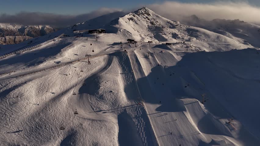 Many Skiing people and tourist on snowy slopes of Cardrona Ski Resort at sunrise. Drone wide shot. Hovering clouds over snowy summit. Ski Lift to base in New Zealand.