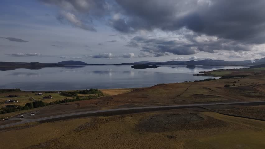 Wide cinematic drone shot over Thingvallavatn Lake in Þingvellir National Park, Iceland. The frame shows a scenic Icelandic landscape with a curving road, calm reflective water, and heavy clouds.