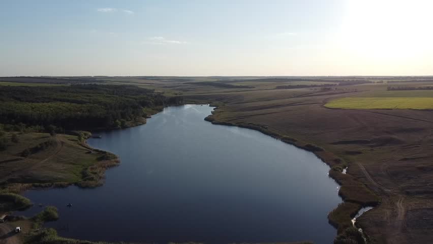 Aerial footage of a calm, dark-blue pond (reservoir) nestled in the wide, green valleys of the Donetsk region, Eastern Ukraine.