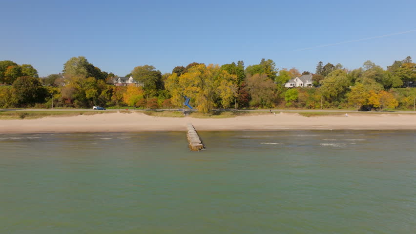 Drone aerial over Lake Michigan on a beautiful fall day, pushing over gentle waves toward the shore, concrete pier, colorful autumn trees, and Sheboygan, Wisconsin neighborhood homes.