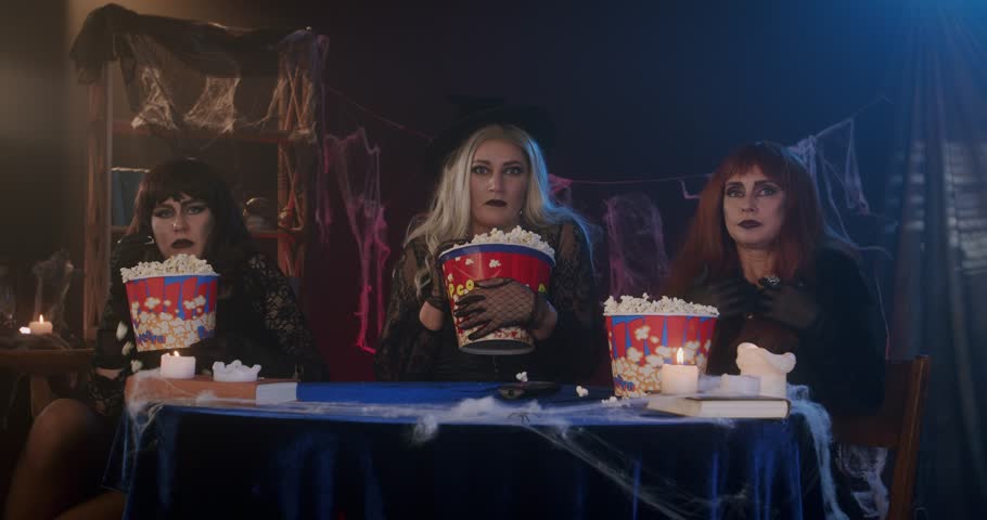 Three women dressed as witches are eating popcorn, watching a movie on TV, and are very excited in a dimly lit, festive Halloween setting with candles. Halloween concept, scary atmosphere, witch party