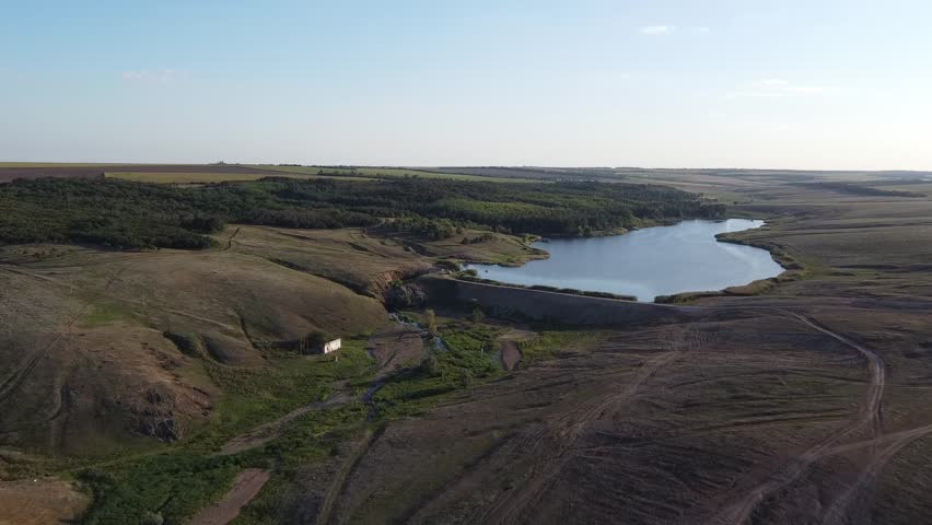 Aerial footage of a calm, dark-blue pond (reservoir) nestled in the wide, green valleys of the Donetsk region, Eastern Ukraine.