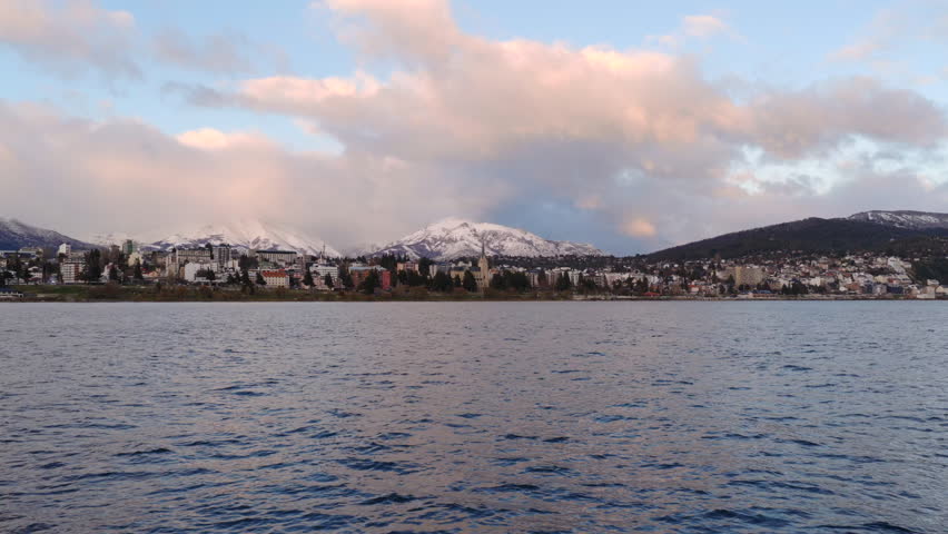 San Carlos de Bariloche city skyline with buildings and snow-capped mountains and Tronador peak reflecting on Nahuel Huapi lake, Patagonia, Argentina, during winter, drone gliding over water
