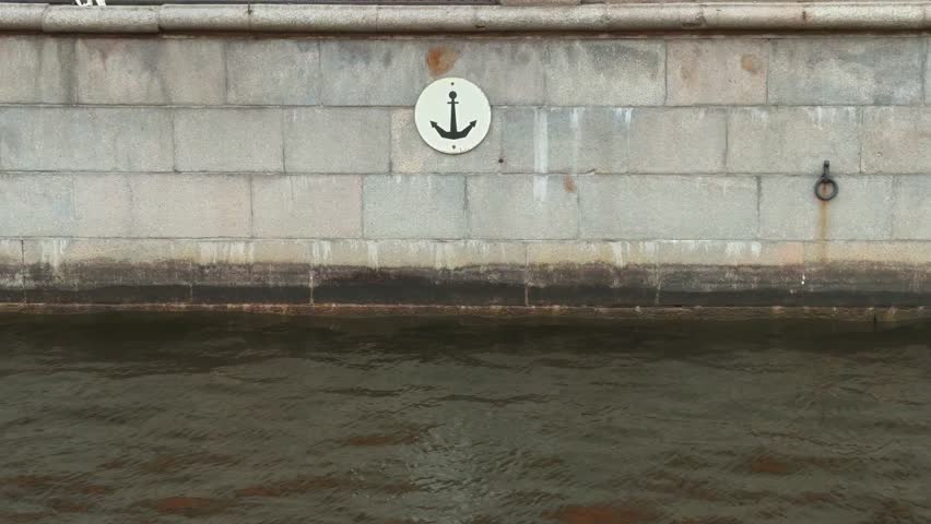 A close-up, straight-on shot of an old, pale-gray, massive granite block quay wall, featuring a white circular sign with a black anchor icon, positioned just above the dark, murky surface of a city