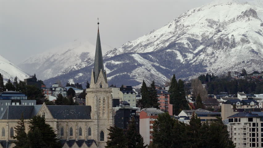 Bariloche cityscape with Cathedral Nuestra Señora del Nahuel Huapi rising among modern buildings, framed by snow-capped Andes peaks under a crisp Patagonian sky, drone parallax shot