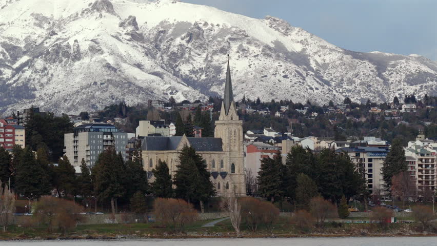 San Carlos de Bariloche cityscape with Bariloche Cathedral by Nahuel Huapi Lake, framed by green trees and snow-capped Andes peaks under a clear blue sky
