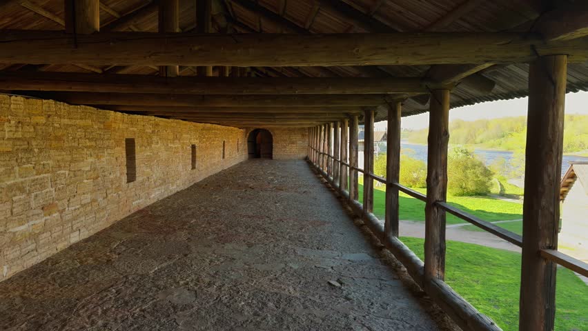 A medium, wide-angle shot of a long, empty covered walkway or gallery running along the inside of a medieval fortress wall made of light-colored, rough stone blocks. The roof and railing are supported