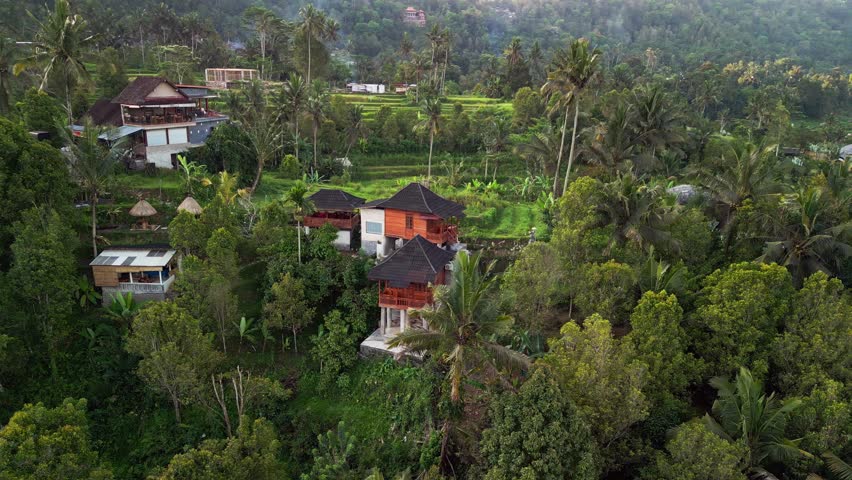 Drone footage of a wooden guest house in the Bali countryside. Overlooks lush rice fields, forested hills, and local villages, showcasing authentic Balinese charm and tranquil tropical landscapes.