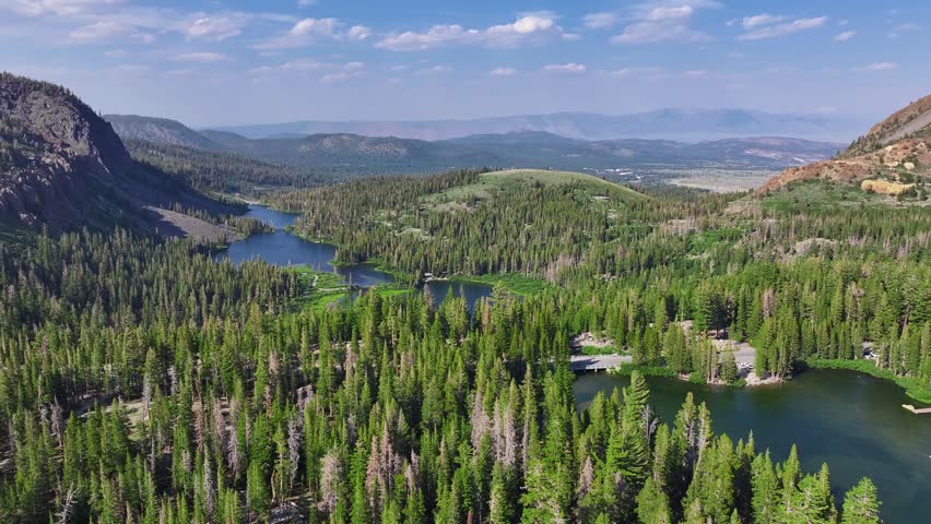 Drone pans across Lake Mary the Mammoth Lakes Basin revealing blue alpine water dense pine forest and red volcanic cliffs with Eastern Sierra peaks beyond. Bright summer daylight smooth lateral motion