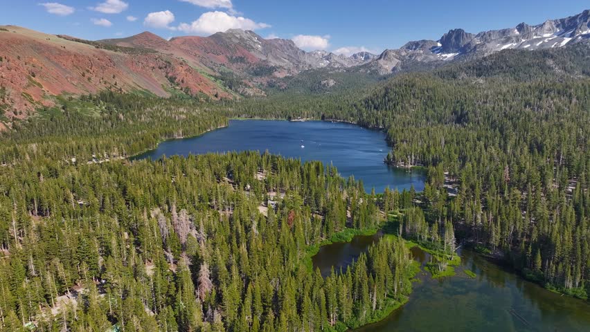 Drone pulls back above Lake Mary in the Mammoth Lakes Basin revealing clear blue water dense pine forest, rugged Eastern Sierra peaks in bright summer light. Expansive alpine scenery and gentle motion
