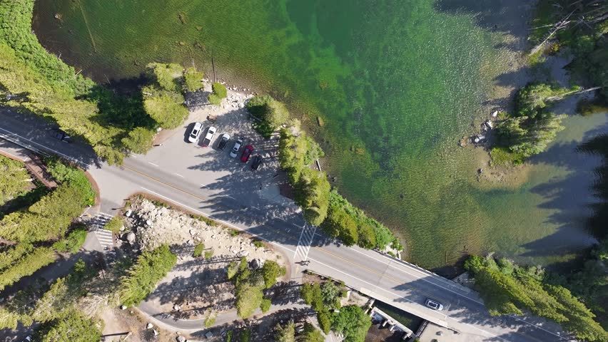 High top down drone pass over the clear green water of Lake Mary in the Mammoth Lakes Basin then drifting to reveal the forested shoreline small dock bridge and parking area in bright sun long shadows