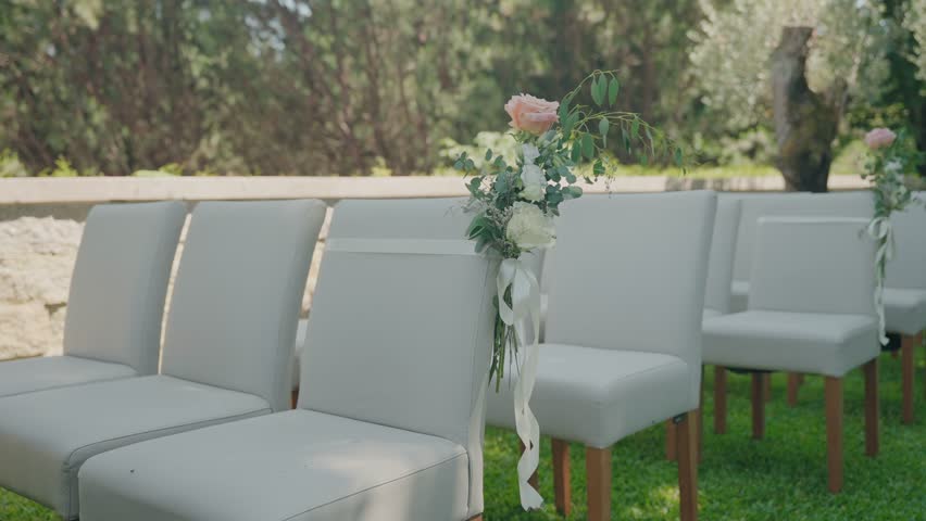 Rows of white chairs arranged on green grass with floral bouquets and ribbons attached for an outdoor wedding setup