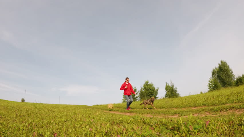 Woman dressed in a vibrant red jacket is walking a dog across a grassy field, surrounded by trees and open sky, capturing a lively outdoor scene filled with joy and connection to nature
