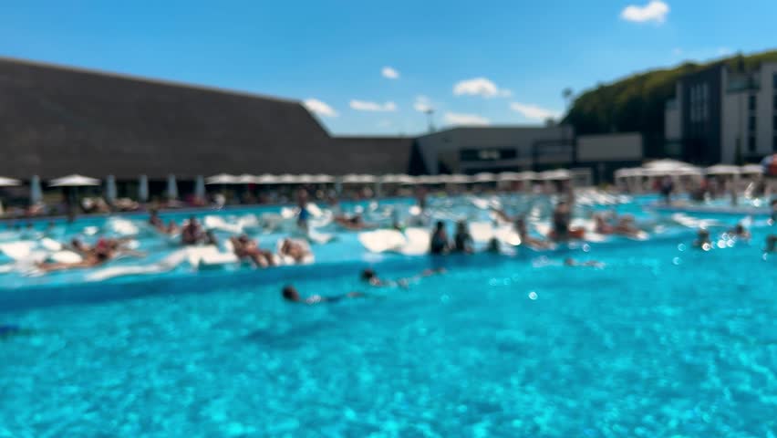 An outdoor pool area. People swim, relax in the water, and sunbathe near the pool under a blue sky on a sunny summer day. Out of focus.