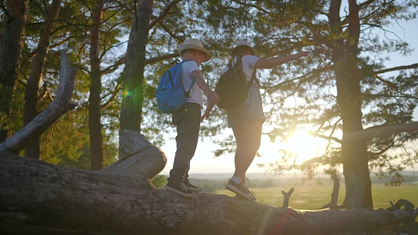 Boy and girl walking on log in forest. Children wearing backpack keep balance on tree. Nature path walking through forest. Girl leads boy carefully. Balance on tree backpack walking children.
