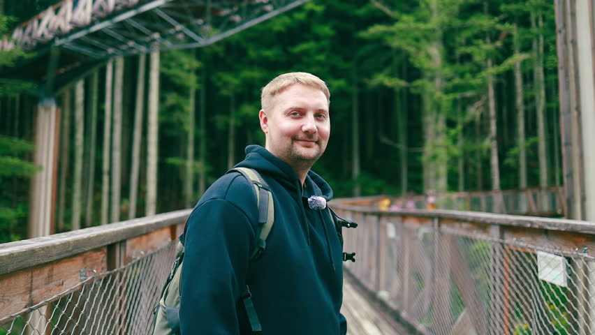 Male traveler with a backpack walking across a wooden bridge in a forest park. Concept of travel, adventure, hiking, exploration, and eco tourism in nature. Scenic outdoor footage showing wanderlust