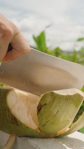 Hands Peeling Outer Shell of Young Coconut with Sharp Cleaver on Balcony under Blue Sky and Green Foliage, close up angle, Tropical Beverage Concept.