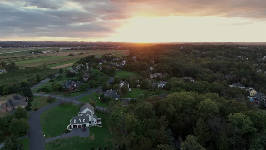 Luxury suburb neighborhood with large houses and swimming pool in garden. Rural farm fields and golden sunrise in distance. Aerial rising wide shot. American countryside city in Pennsylvania.