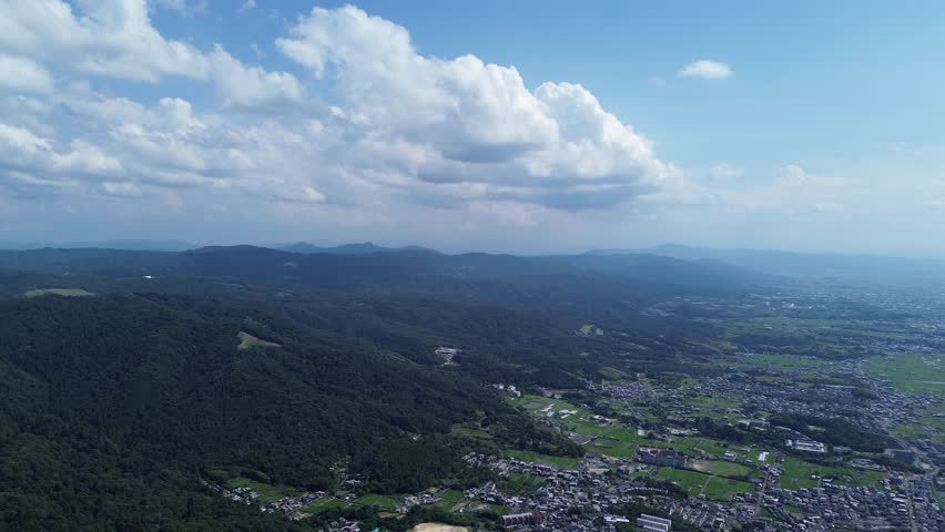 Summer aerial scene of Gokoku Shrine in Nara, Japan, showcasing the shrine amidst a blend of natural greenery and urban elements under a vivid blue sky with fluffy clouds.