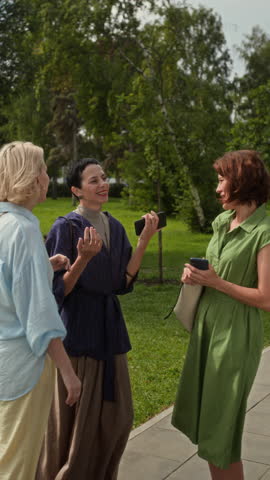 Vertical shot of three middle-aged Caucasian women standing outdoors on summer day, holding smartphones in hands and chatting