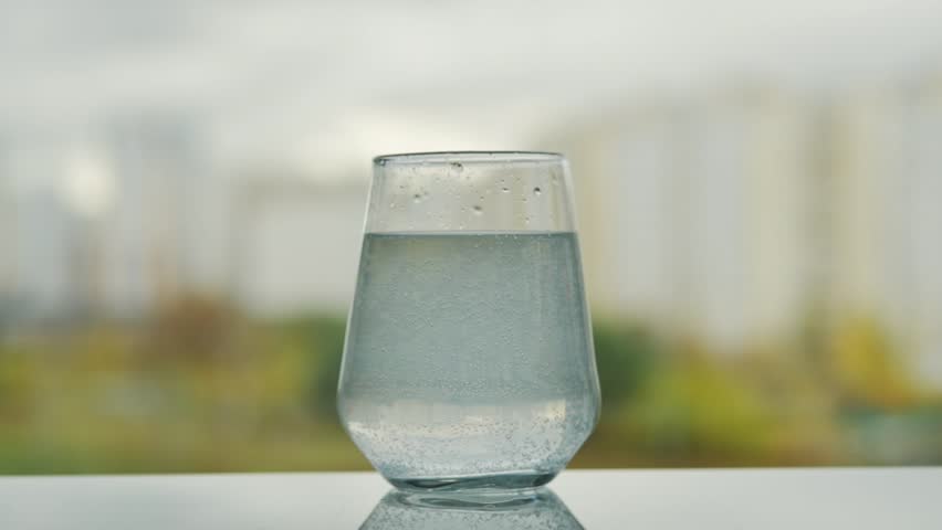 Hand reaches for glass with dissolving tablet, sparkling bubbles in water, soft cityscape in background, daily health care concept.