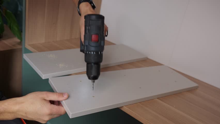 Worker using electric drill to drilling hole in wooden board. Man assembling furniture at home, preparing shelf to installing in self made cabinet. Concept of renovation and interior improvement