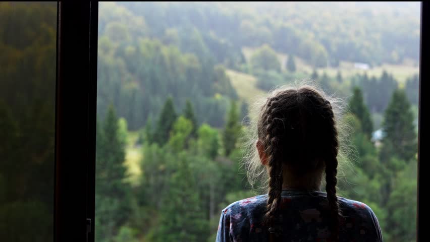 Girl looking through window at mountain landscape. Child gazing through window with scenic mountains and forest outside. Peaceful family travel moment.