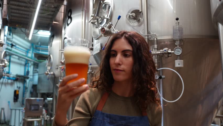 Female brewmaster examining and tasting a glass of freshly brewed craft beer. Her quality control process inside a modern brewhouse
