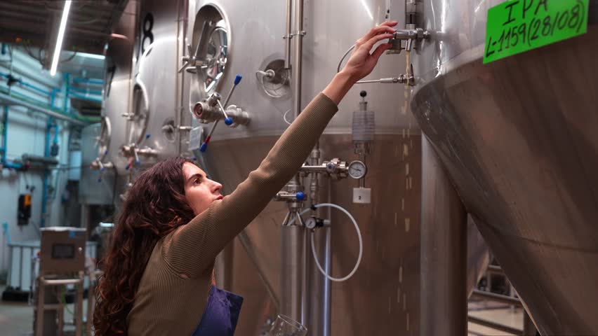 Young woman pouring a fresh glass of beer from a fermentation tank in a brewery. Professional brewmaster checking the quality of her product