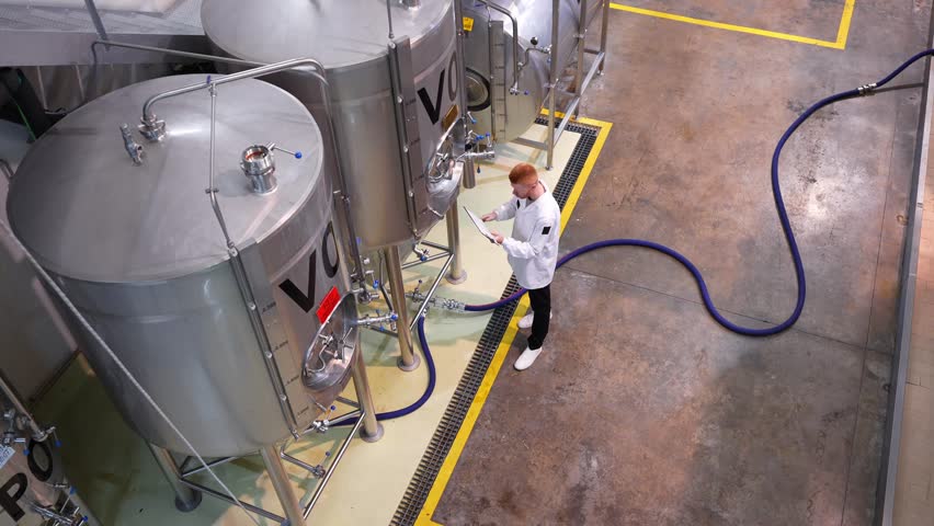 High angle view of a brewmaster monitoring the brewing process. The specialist is writing on a clipboard while checking the machinery