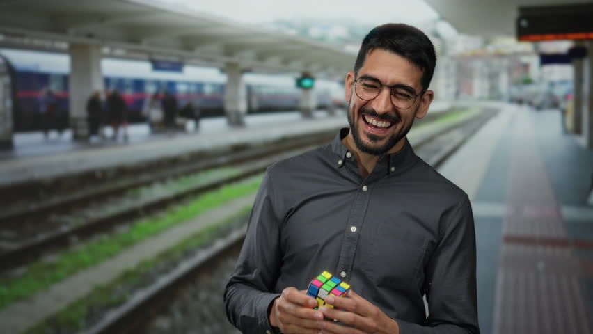 Young man smiling while holding a colorful puzzle cube at a train station, showcasing joy and engagement in a lively outdoor railway environment.