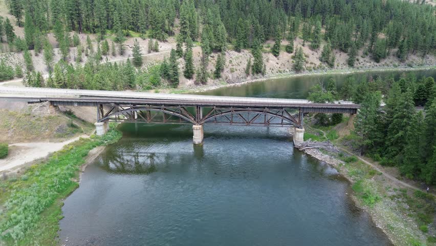 Railroad bridge over Clark Fork River in Montana. Unusual design is due to additional reinforcing structure to carry heavier trains. Flyover shot goes up river.