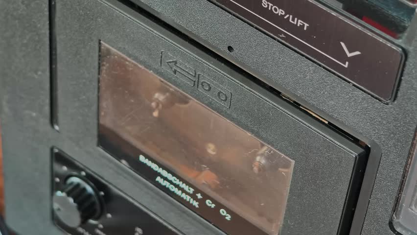 Angled shot of hand opening tape deck door, inserting vintage yellow cassette, and closing it. Shows internal player mechanism, pinch rollers, and German text on the black textured housing of the