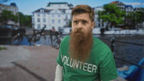 Man with long red beard wearing volunteer shirt raises fist on street by canal in urban city; solidarity support activism. - Powered by Shutterstock - Get 15% off with code: PIKWIZARD15