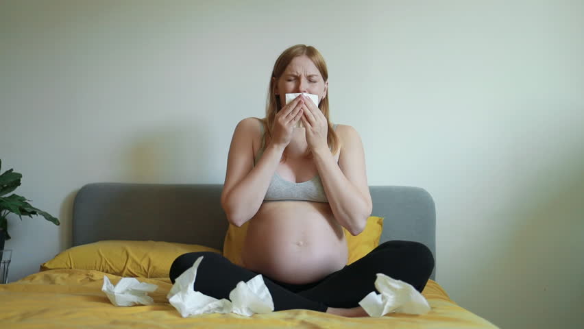 Video of a pregnant woman sitting on bed and using a tissue, suffering from cold or allergy. Concept of pregnancy health, home care, illness, and seasonal sickness.