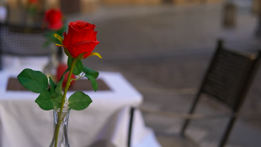 Red rose in vase adorning outdoor restaurant table. Delicate red rose standing elegantly in glass vase, gracing rustic restaurant table with soft-focused european street scene