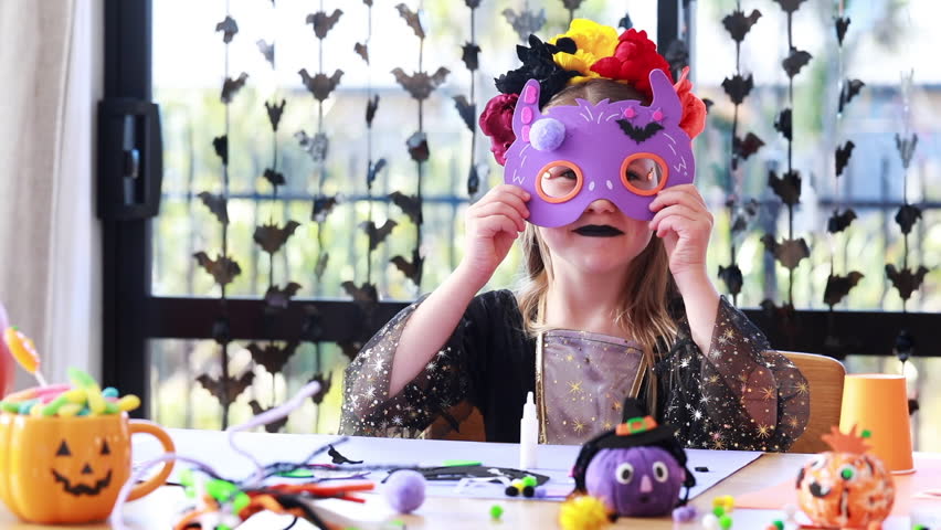 Child in halloween costume crafts colorful mask at creative arts table. Concept of Halloween crafts for kids.