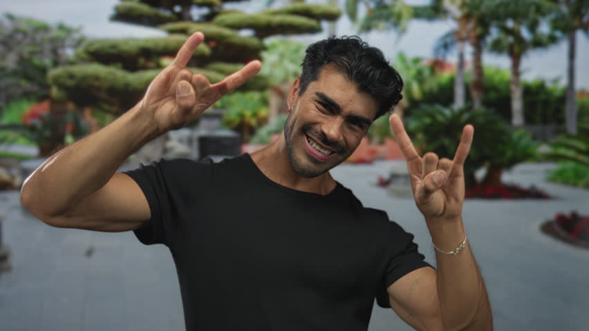 Young hispanic man making rock horns gesture with hands and smiling broadly on street beside green trees and fountain; rock metal fun.