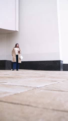 brunette woman wearing coat walking in the street along the large building, minimal urban background using smartphone