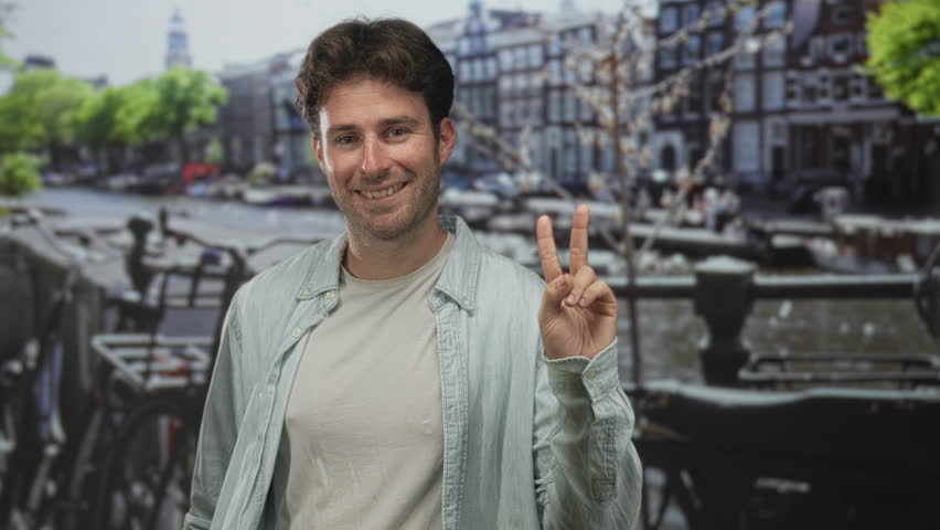 Man showing peace sign with hand on street by amsterdam canal, smiling and relaxed; cheerful travel.