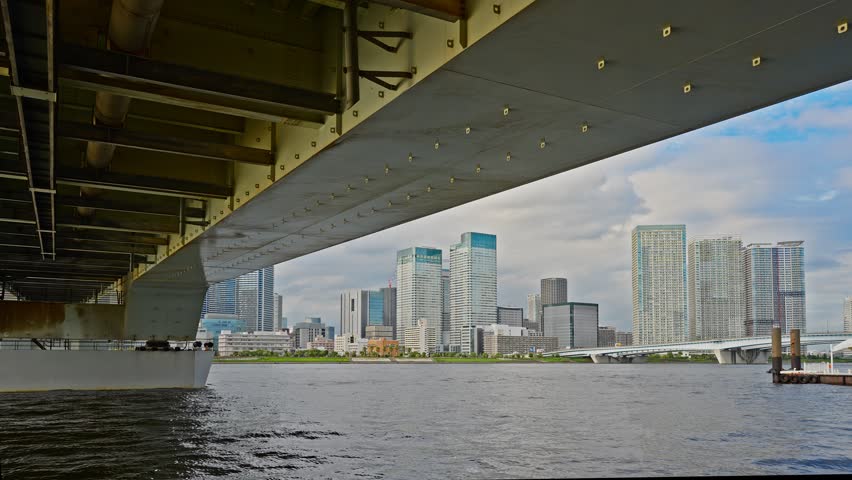 A low-angle view of the modern Tokyo skyline seen from directly underneath the structure of a large urban bridge.