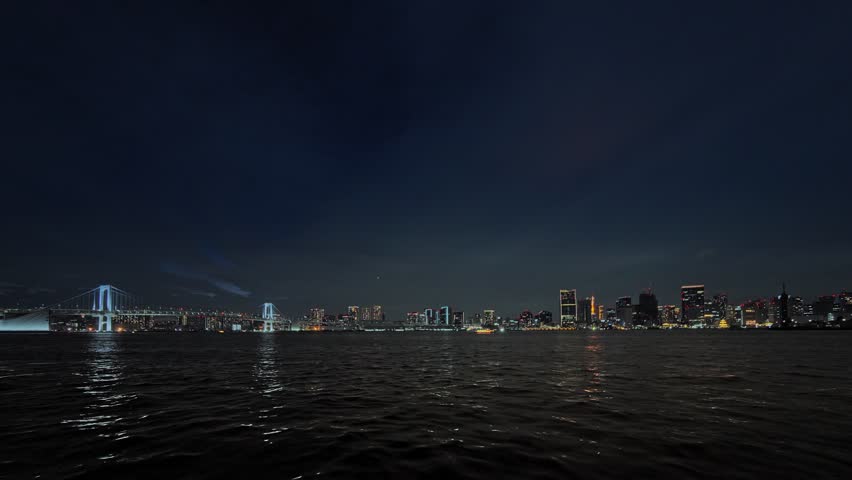 A very wide, dark night shot of a metropolitan skyline with a large, distant suspension bridge (likely the Rainbow Bridge).