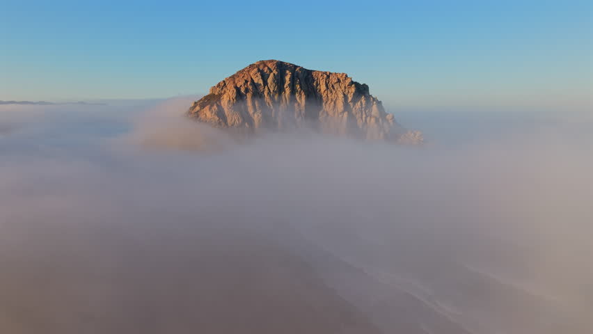 Cinematic aerial view of Morro Bay rock formation rising above the clouds at sunrise, showcasing natures beauty, perfect for adventurers seeking unforgettable experiences. California, USA