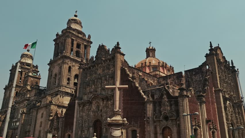 Close-up view of the ornate baroque façade and towers of the Mexico City Metropolitan Cathedral, with the Mexican flag waving in the background under a clear sky.